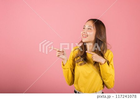 Portrait Asian beautiful young woman smiling standing pointing finger out on side pink background, Thai happy face excited female point into empty with copy space for text, point side away 117241078