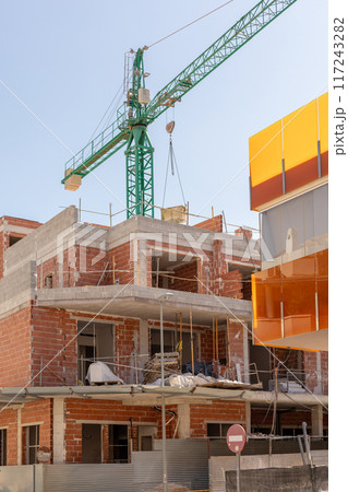 Vivid capture of construction site showcasing building in progress with brickwork and crane lifting materials. Ideal for real estate, development projects, and construction industry themes. 117243282