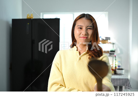 Playful young asian woman holding a spatula standing in the kitchen. Domestic life and cooking concept 117245925