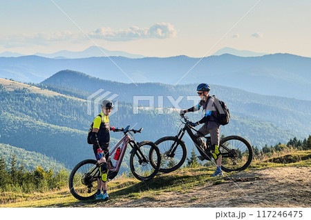 Two cyclists men riding electric bikes outdoors. Smiling male tourists resting on the top of hill, enjoying beautiful mountain landscape, wearing helmet and backpack. Concept of active leisure. 117246475