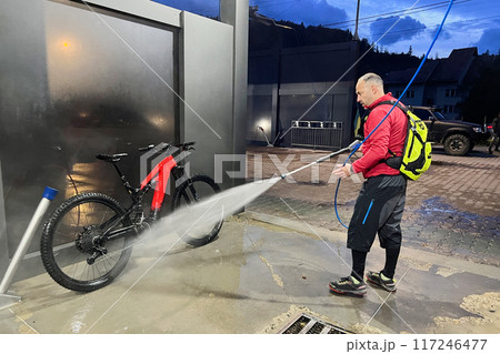 Cyclist using high-pressure water spray to clean electric mountain bike on outdoor washing station. 117246477