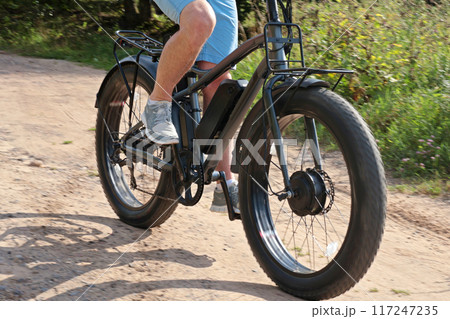 Cyclist on electric bicycle riding along path in field in countryside at sunset close-up. Sport and health. Summer hobby. Cyclist on electric bicycle riding along path in field in countryside at sunset close-up. Sport and health. Summer hobby. 117247235
