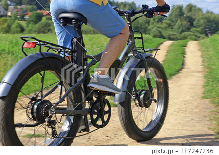 Cyclist on electric bicycle riding along path in field in countryside at sunset close-up. Sport and health. Summer hobby. Cyclist on electric bicycle riding along path in field in countryside at sunset close-up. Sport and health. Summer hobby. 117247236