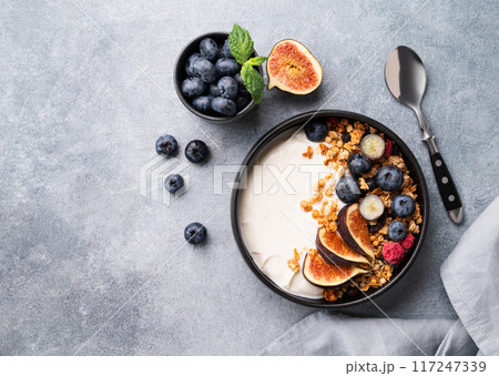 Natural yogurt with muesli, berries and figs in a black bowl on a blue background with napkin. Natural yogurt with muesli, berries and figs in a black bowl on a blue background with napkin. 117247339
