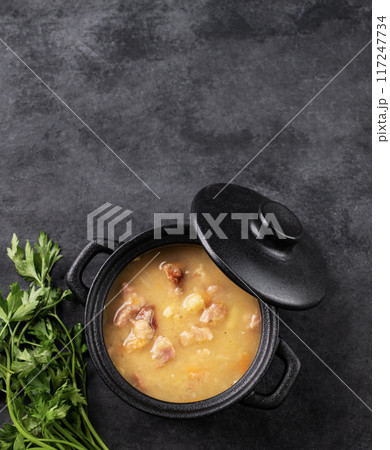 Pea soup with smoked ham in black soucepan on dark background with herb. 117247734