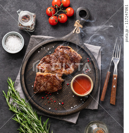 BBQ beef steak grill with smoke, sauce, tomatoes, salt and rosemary on black wooden board BBQ beef steak grill with smoke, sauce, tomatoes, salt and rosemary on black wooden board 117247861