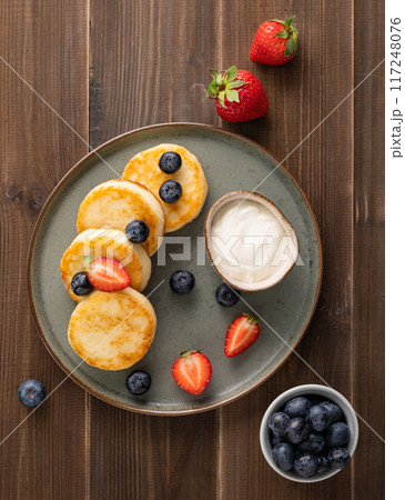 Cottage cheese pancakes with sour cream, blueberries and strawberries on a dark wooden background.  117248076