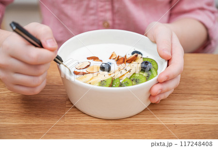 The hands of a girl in a pink shirt hold a bowl of yogurt, granola and fresh fruit on a wooden table 117248408