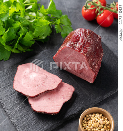 Seasoned smoked beef with slices on black slate on dark background with fresh vegetables and herb. Seasoned smoked beef with slices on black slate on dark background with fresh vegetables and herb. 117248427