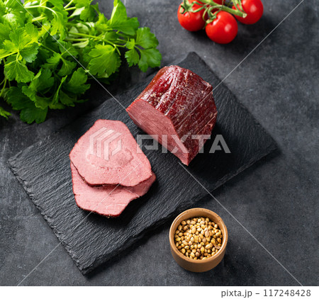 Juicy baked roast beef on a slate board with tomatoes and fresh herbs on a dark  background. 117248428