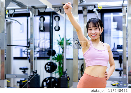 スポーツジムでポージングする笑顔の若い女性　ダイエット　パーソナルジム　筋トレ女子　ガッツポーズ 117248451