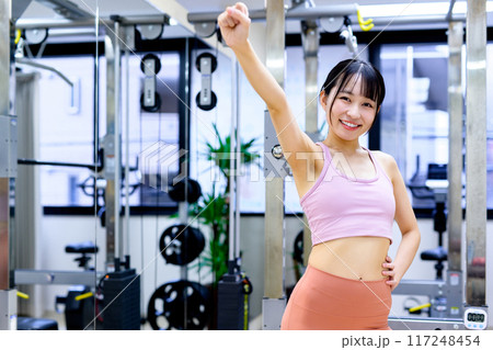 スポーツジムでポージングする笑顔の若い女性　ダイエット　パーソナルジム　筋トレ女子　ガッツポーズ 117248454