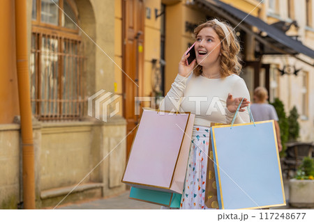 Excited woman shopaholic consumer after shopping sale with full gift bags talking on smartphone Excited woman shopaholic consumer after shopping sale with full gift bags talking on smartphone 117248677