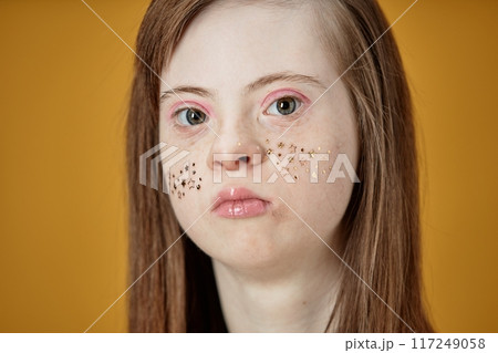 Portrait of young woman with Down Syndrome, looking straight at the camera Her face adorned with star stickers, hair straight and long, against plain background Portrait of young woman with Down Syndrome, looking straight at the camera Her face adorned with star stickers, hair straight and long, against plain background 117249058