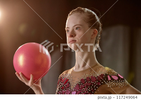 Portrait of girl with Down syndrome holding a shiny pink ball, facing camera under studio lighting, wearing decorative costume with colorful patterns 117249064
