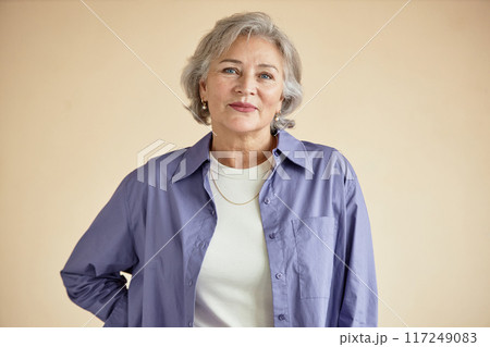 Portrait of an elderly Caucasian woman smiling while wearing lavender shirt and white top. She is standing against neutral background with relaxed posture and friendly expression Portrait of an elderly Caucasian woman smiling while wearing lavender shirt and white top. She is standing against neutral background with relaxed posture and friendly expression 117249083