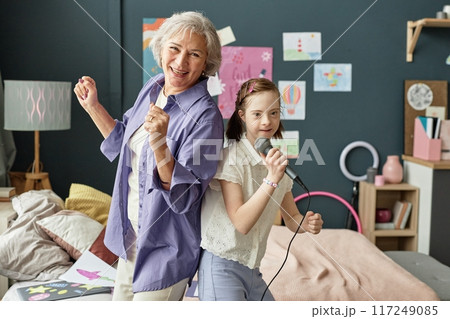 Elderly woman and young girl singing into microphone in cozy bedroom surrounded by drawings and artworks creating joyful atmosphere 117249085