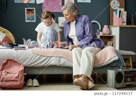 Senior woman assisting young girl with school assignments while sitting in cozy bedroom setting. Intergenerational bonding between grandmother and granddaughter without any distractions 117249117