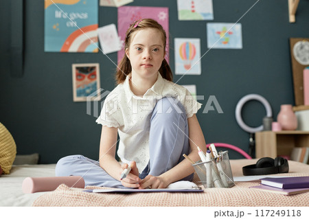 Portrait of a young girl with Down syndrome sitting on bed while drawing on paper in cozy bedroom setting 117249118