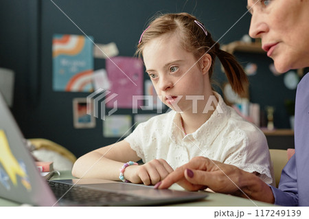 Girl with Down Syndrome engaging in home learning on laptop with adult supervision in background, focusing on screen and participating in educational activity Girl with Down Syndrome engaging in home learning on laptop with adult supervision in background, focusing on screen and participating in educational activity 117249139
