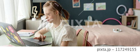 Side view of young girl with Down syndrome using laptop while studying online at home, side view with copy space 117249155