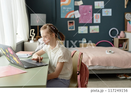 Young girl with Down syndrome sitting at desk in bedroom using laptop for online studies Engaging in learning surrounded by cozy decor Young girl with Down syndrome sitting at desk in bedroom using laptop for online studies Engaging in learning surrounded by cozy decor 117249156