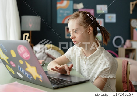 Girl using laptop in cozy room decorated with artwork and a lamp, focusing on screen and working diligently while sitting at desk with curtain letting in light without casting shadows 117249157