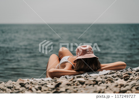 A woman is laying on the beach with a pink hat on her head. She is wearing a white bikini and is laying on a towel. The beach is rocky and the water is calm. Scene is relaxed and peaceful. 117249727