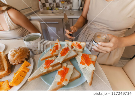 A woman is cutting bread and putting jelly on it. The table is set with a variety of food items, including sandwiches, fruit, and a jar of jelly. The woman is wearing a dress. A woman is cutting bread and putting jelly on it. The table is set with a variety of food items, including sandwiches, fruit, and a jar of jelly. The woman is wearing a dress. 117249734