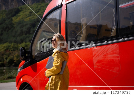 A woman in a yellow jacket stands next to a red bus A woman in a yellow jacket stands next to a red bus 117249753