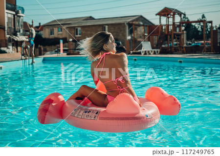 A woman is sitting on a pink flamingo float in a pool. The scene is bright and cheerful, with the woman enjoying her time in the water. 117249765