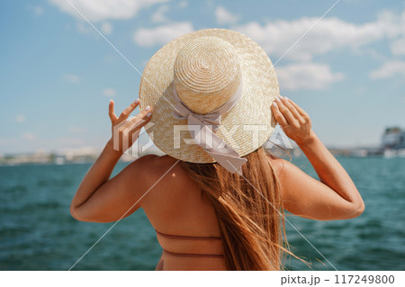 A woman in a swimsuit sits with her back holding a hat, looks at the ocean, sunny day, relaxes. A woman in a swimsuit sits with her back holding a hat, looks at the ocean, sunny day, relaxes. 117249800