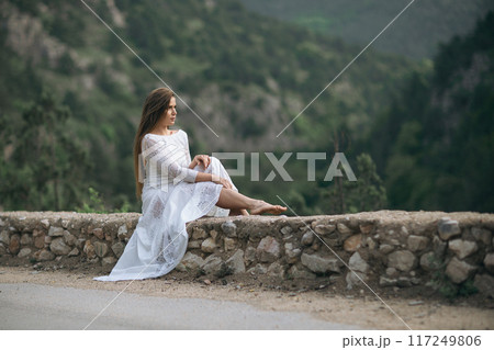 A woman in a white dress is sitting on a stone wall. The scene is serene and peaceful, with the woman looking out into the distance. The dress is long and flowing, adding to the sense of calmness. A woman in a white dress is sitting on a stone wall. The scene is serene and peaceful, with the woman looking out into the distance. The dress is long and flowing, adding to the sense of calmness. 117249806