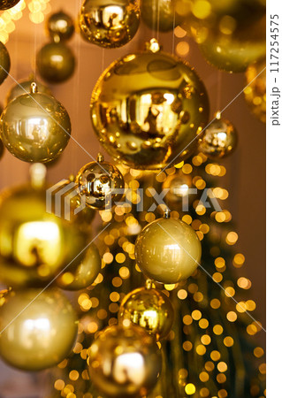 Many large golden Christmas balls look hanging against the background of the Christmas interior. Festive background in golden awards, bokeh, selective focus Many large golden Christmas balls look hanging against the background of the Christmas interior. Festive background in golden awards, bokeh, selective focus 117254575