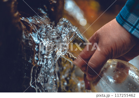 A hand brings a transparent bottle to a spring of drinking water. A man collects mineral water from a spring in the autumn forest. The traveler stopped at a spring to drink and collect clean water. 117255462
