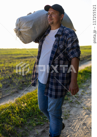 A tired worker returns from the field with a sack over his shoulder at sunset. A man walks along the field after a hard day's work. Harvesting concept. A tired worker returns from the field with a sack over his shoulder at sunset. A man walks along the field after a hard day's work. Harvesting concept. 117255521