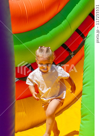 Having rolled down from an inflatable bright slide on a trampoline, the child runs to play to be daring. In summer, a child on an inflatable trampoline. There is a colorful trampoline in the park. Having rolled down from an inflatable bright slide on a trampoline, the child runs to play to be daring. In summer, a child on an inflatable trampoline. There is a colorful trampoline in the park. 117255531
