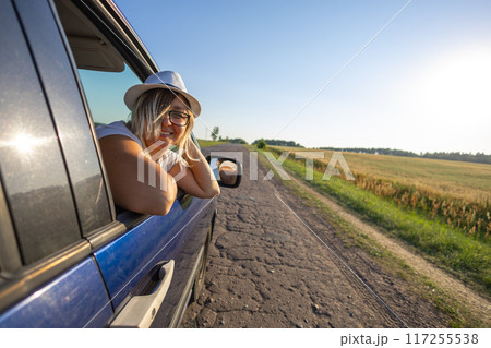 The wind blows the woman's hair in the car window. Summer travel by car. A woman looks out the car window at a beautiful sunset against the blue sky. The wind blows the woman's hair in the car window. Summer travel by car. A woman looks out the car window at a beautiful sunset against the blue sky. 117255538