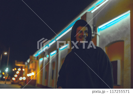 On the street of a city at night, a handsome man laughs. A man in a hood smiles sweetly at night in the neon light of the city. City nightlife concept. 117255571