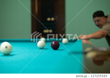 A man aims a cue at a billiard ball on a green table. A man hits a billiard ball and tries to pocket it. A man plays Russian billiards with his friends. Concept of a man playing billiards. 117255583