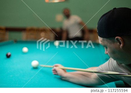 A group of friends plays billiards. The man hits the ball with a cue and hammers the meat into the pocket. People have fun playing billiards in the evening. Concept of a company playing billiards. 117255594