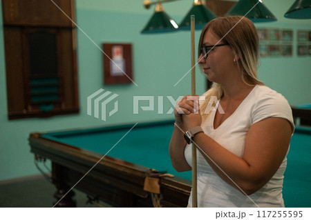 Copy space. A beautiful woman holding a billiard cue in her hands looks to the side, thinking about the next shot. A woman enjoys playing billiards in her free time. Copy space. A beautiful woman holding a billiard cue in her hands looks to the side, thinking about the next shot. A woman enjoys playing billiards in her free time. 117255595