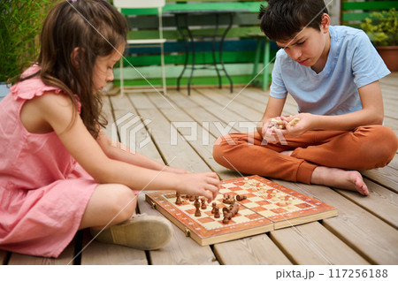 Two kids playing chess on a wooden deck outdoors on a sunny day 117256188