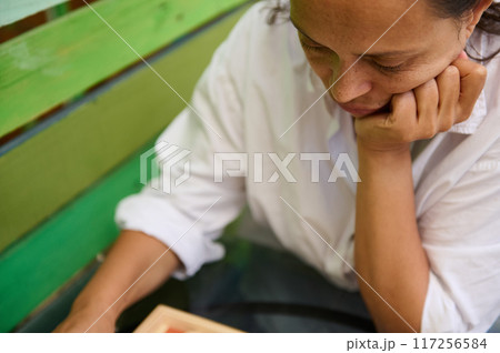 Woman deep in thought sitting on a green bench wearing a white shirt outdoors 117256584