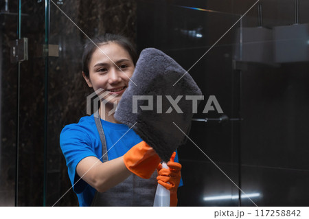 Beautiful housewife wearing orange cleaning gloves uses spray and cloth to clean the clear glass in the room. Beautiful housewife wearing orange cleaning gloves uses spray and cloth to clean the clear glass in the room. 117258842