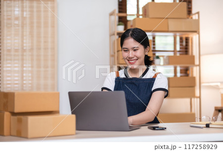 Young woman, small business owner selling products online, accepting online product orders via laptop, sits in a room with boxes of products. 117258929