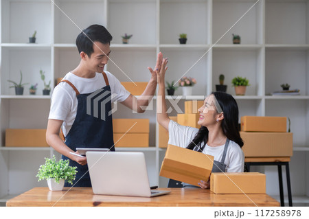 Two online sellers celebrate together in a room full of merchandise boxes as they meet their sales goals. 117258978