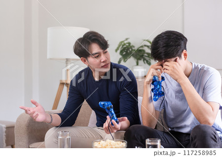A lgbt male couple sits together playing a pc game with a joystick console and expresses their sorrow over a loss. 117258985