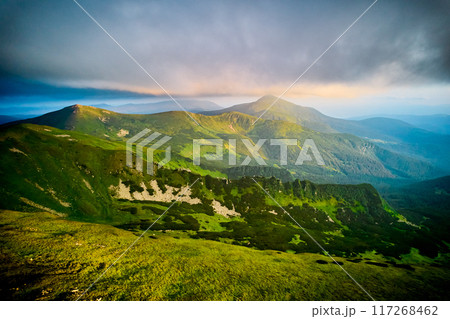 Breathtaking aerial view of green, rugged mountain peaks under dramatic sky. Scene captures contrast between vibrant landscape and moody clouds, with sunlight breaking through. Carpathians, Chornogora 117268462