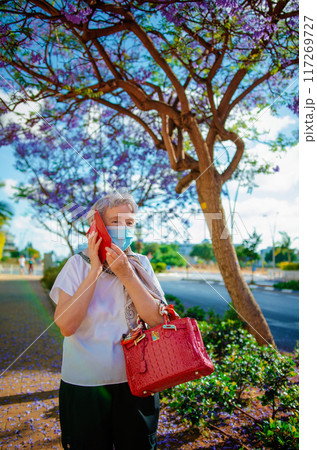 Fashionable Senior Woman with Red Handbag and Phone Case in Protective Mask Talks on Phone by Jacaranda Tree. 117269727